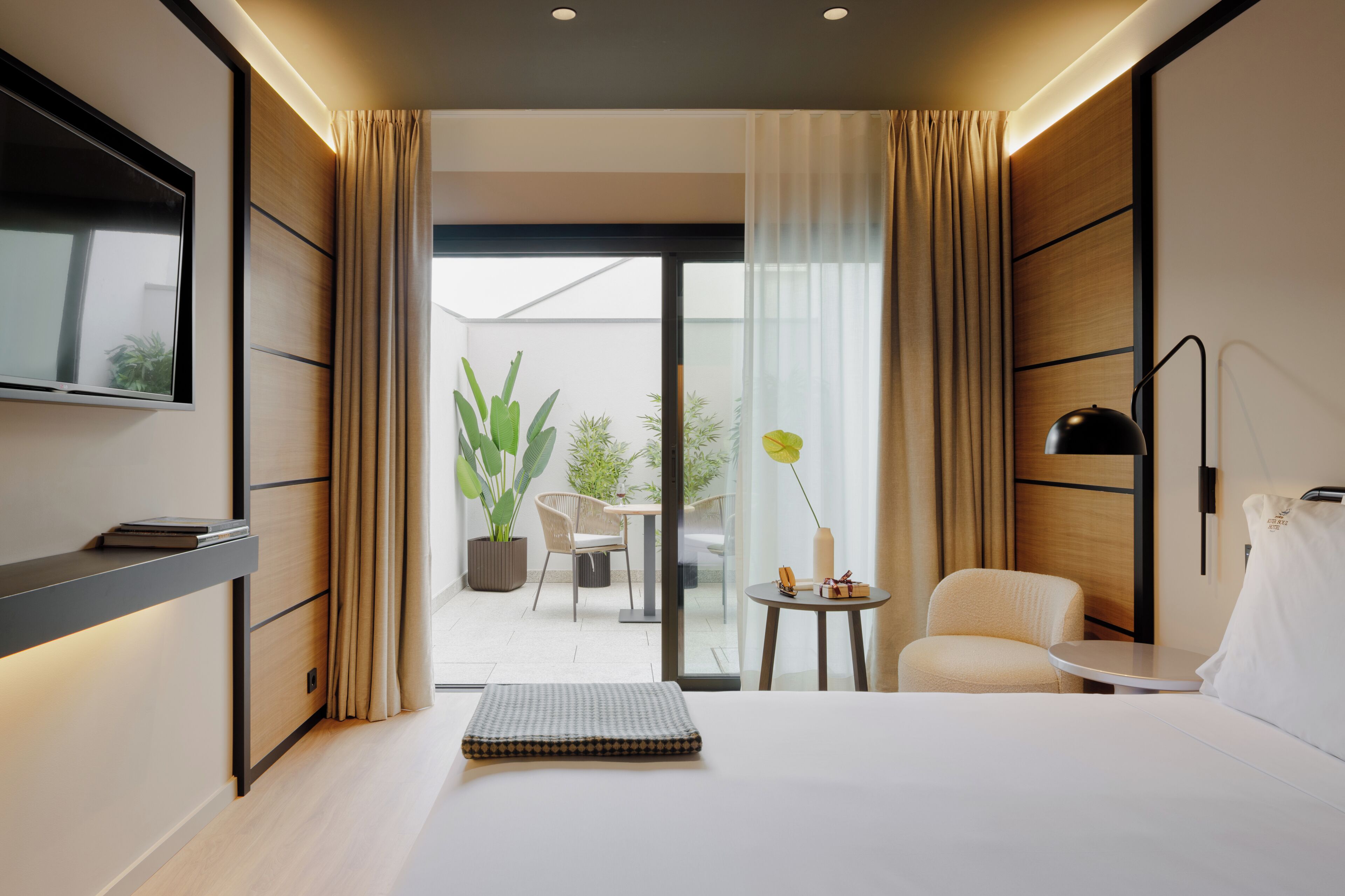 A modern hotel room with a bed, a TV mounted on the wall, a small table with a lamp, and a view of the outdoor patio with plants and a chair.