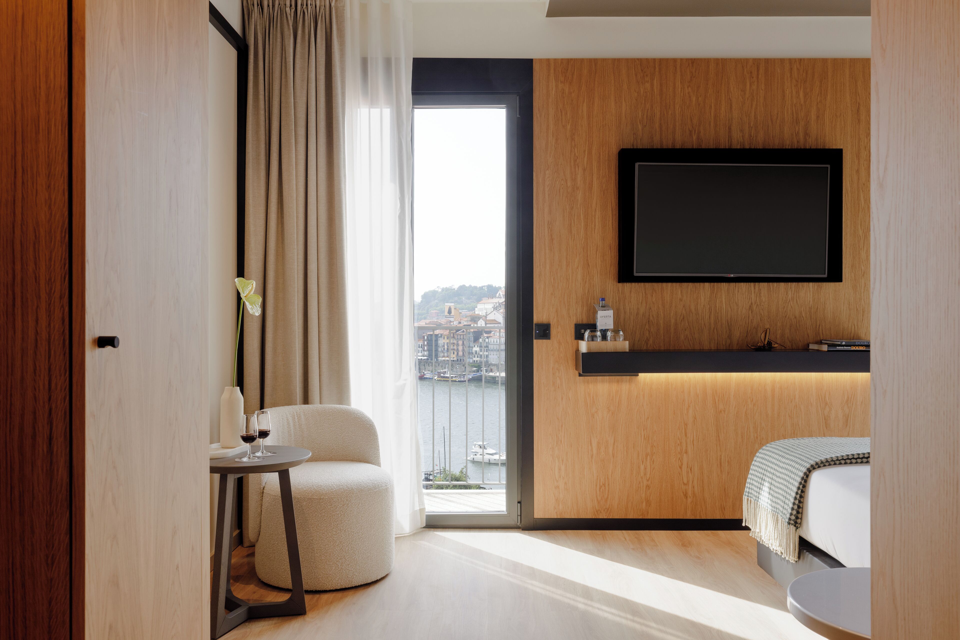Une chambre d’hôtel moderne dotée d’une grande fenêtre, d’un téléviseur à écran plat fixé au mur, d’une petite table ronde agrémentée d’un vase et de verres à vin, et d’un lit confortable offrant une vue sur la ville.