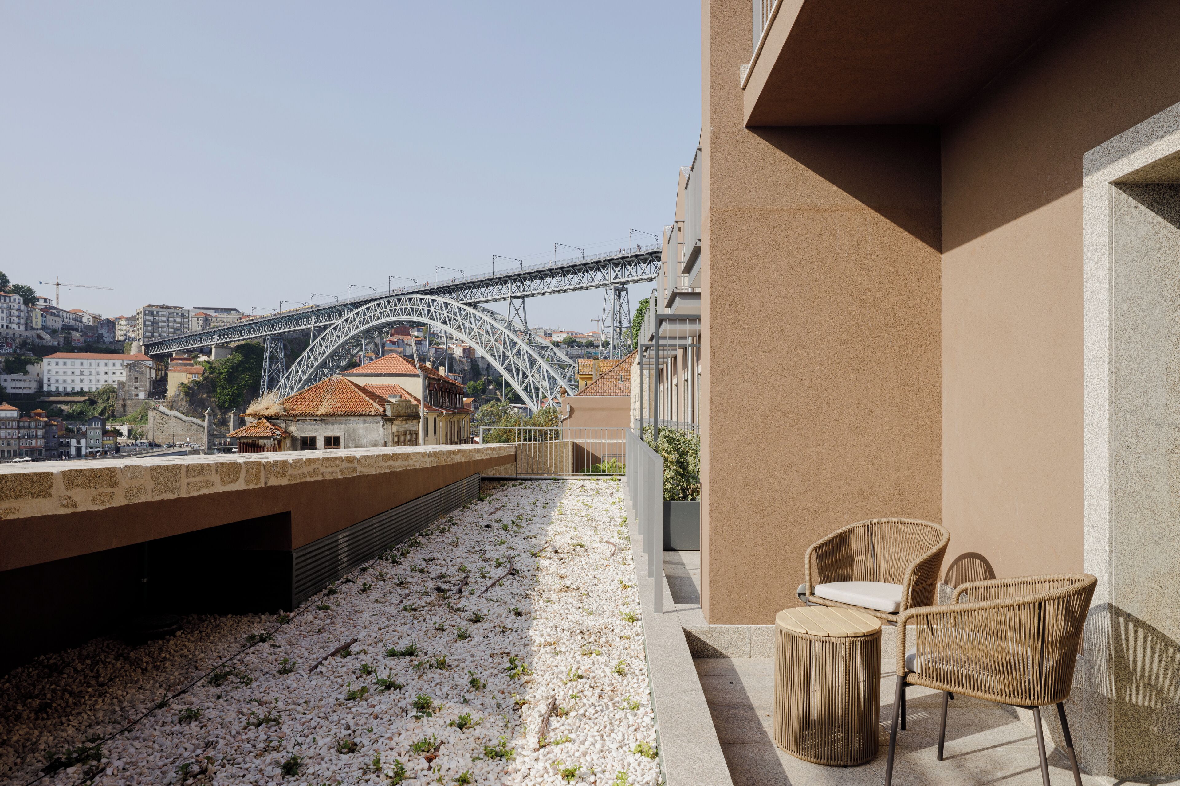 A rooftop terrace with a view of a bridge, wicker furniture, and a pebble-covered ground.