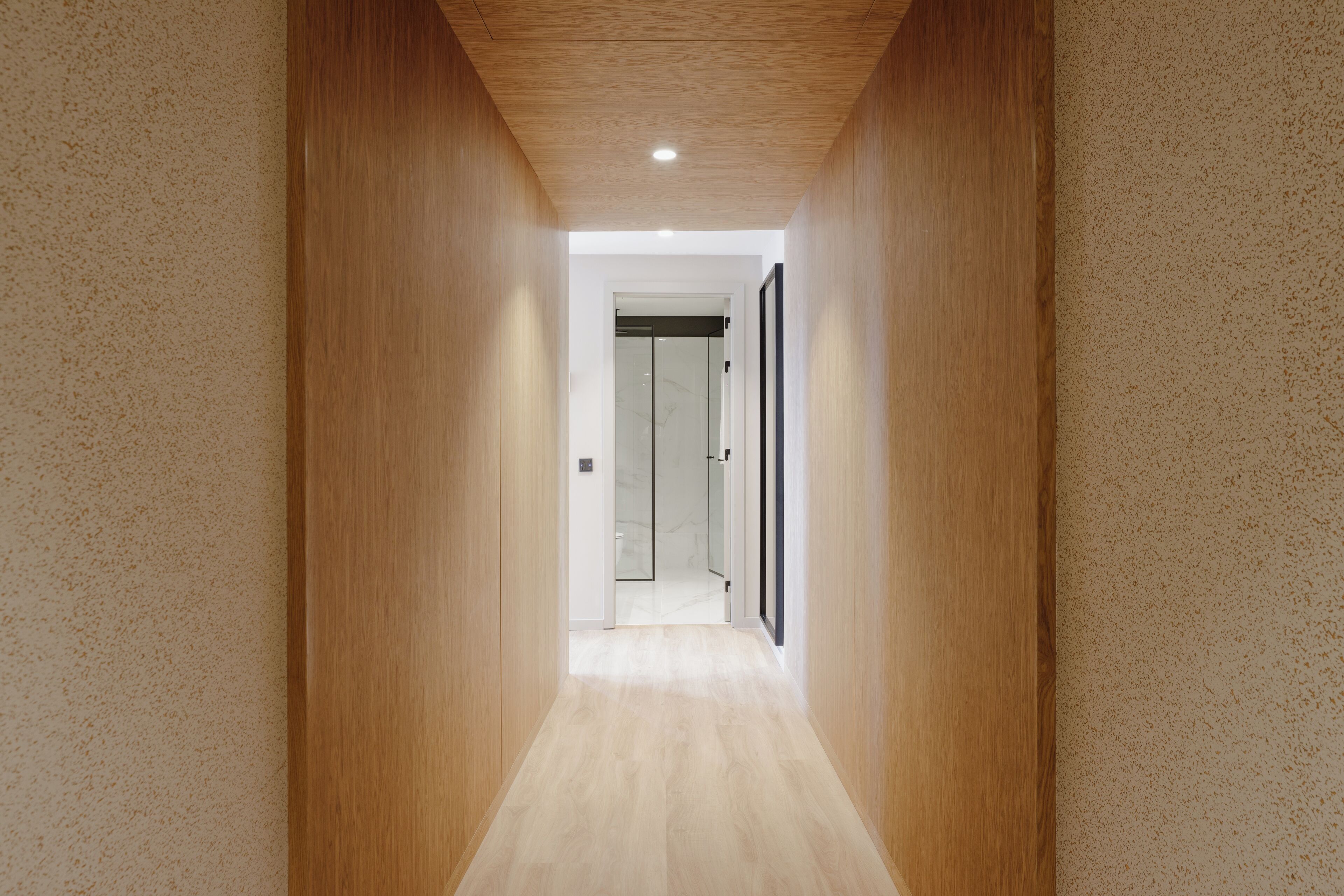 A narrow hallway with wooden walls and a light wooden floor, leading to a glass door.
