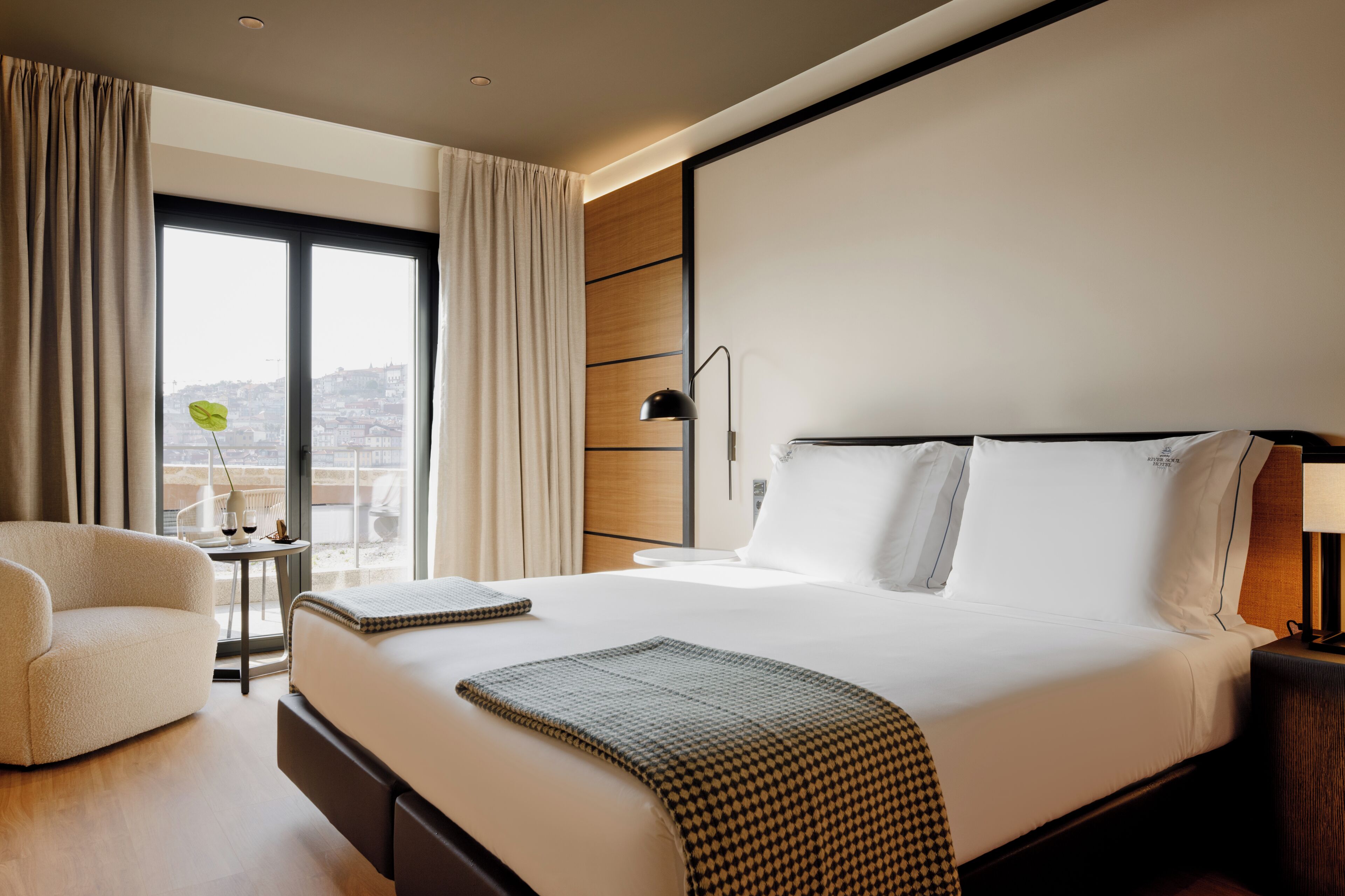 A hotel room with a large bed, a chair, a small table with a plant, and a view of the city through the window.