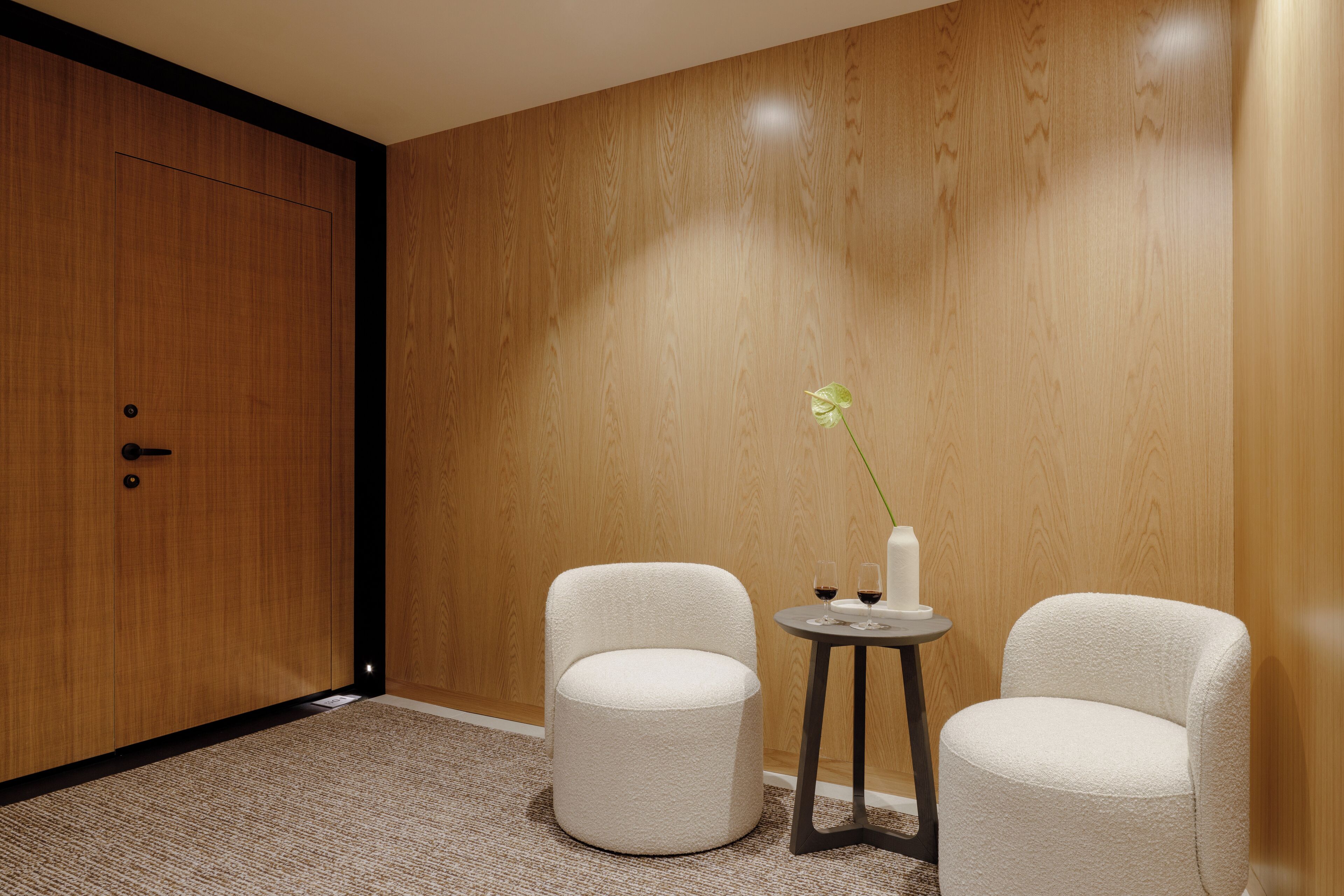 Una habitación con una pared de madera, una puerta, dos sillas blancas, una mesita redonda con un florero y dos vasos.