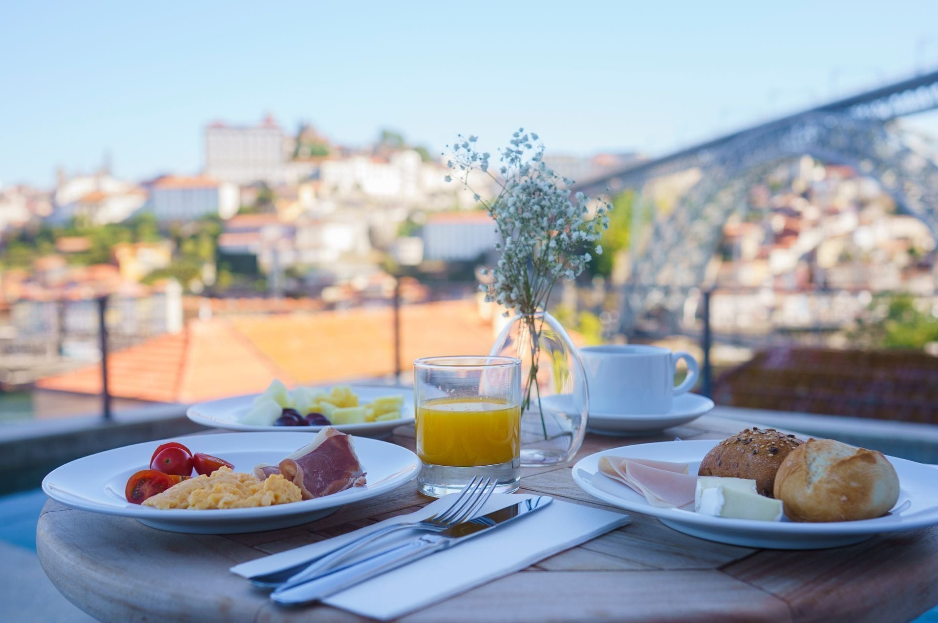 Ein Frühstückstisch mit Tellern mit Eiern, Schinken, Käse und Brot, ein Glas Orangensaft und eine Tasse Kaffee, mit Blick auf eine Brücke und Gebäude im Hintergrund.