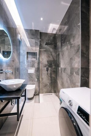 A modern bathroom with a large mirror, a white sink, a shower, and a washing machine. - Day LUX Premium Apartments (Sarandë)