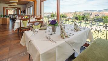A restaurant with tables set for dining, a balcony with a view of the city, and indoor seating with wooden floors.