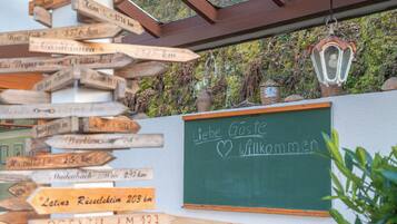 Un poste de madera con varias indicaciones de distancia, una pizarra con la frase 'Herzlich Willkommen' escrita y una mesa con sillas en un entorno exterior.