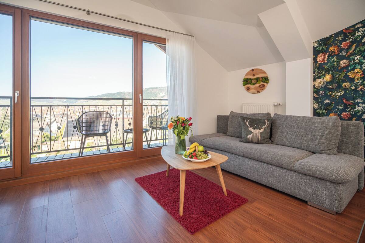 Ein Wohnzimmer mit einem grauen Sofa, einem kleinen Holztisch mit Obst und einem roten Teppich.