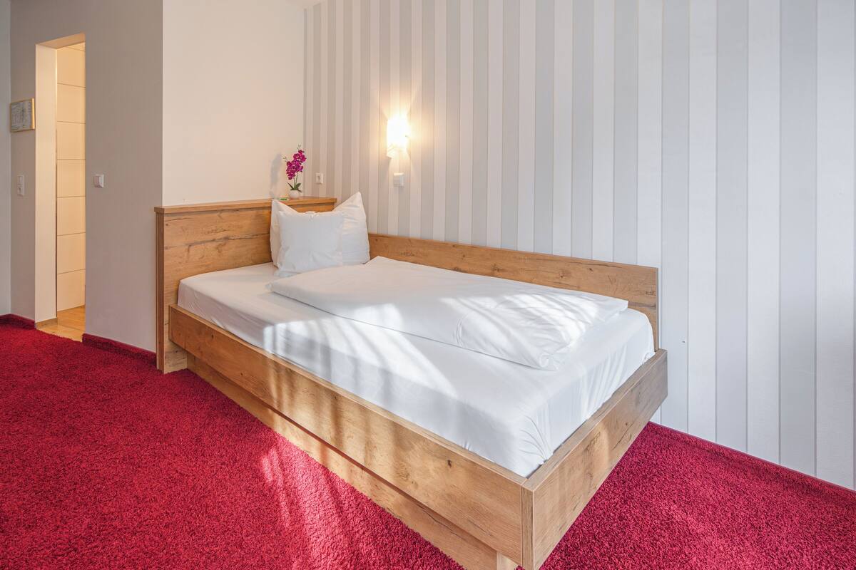 Ein Zimmer mit einem hölzernen Bett, weißer Bettwäsche, einem roten Teppich, einer Wandleuchte und einer Tür.