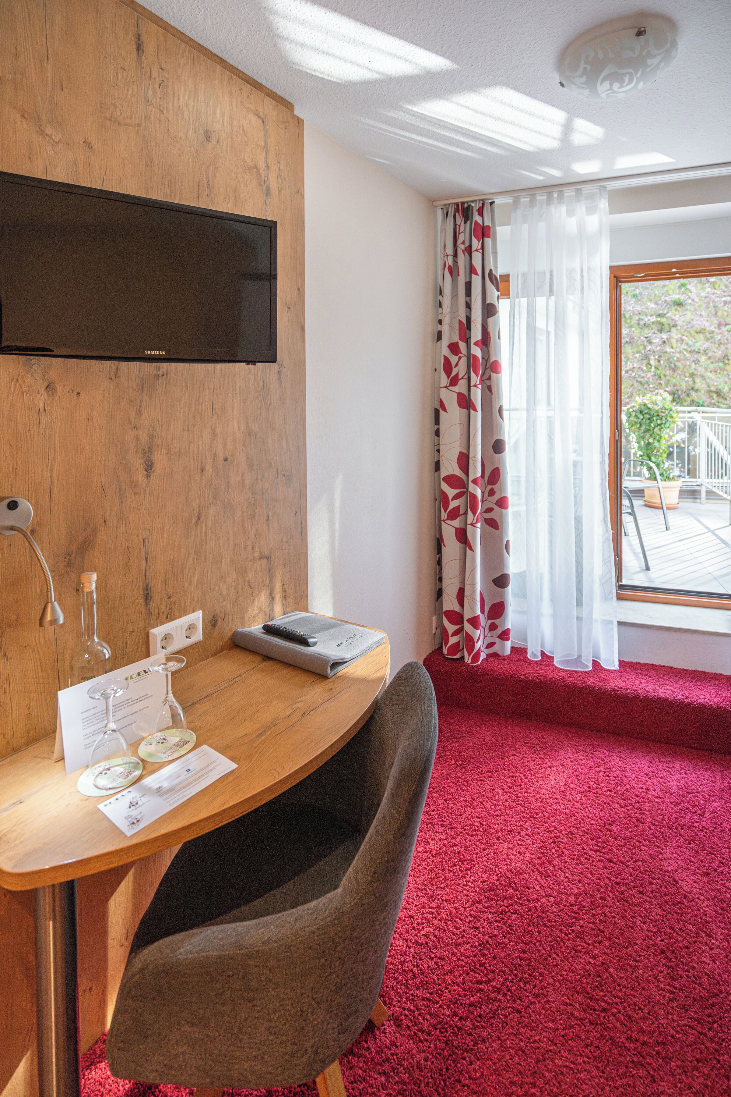 A room with a wooden desk, a flat-screen TV, a red carpet, and a window with curtains.