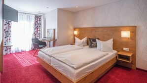 A hotel room with a wooden bed, red carpet, white bedding, a desk with a chair, and a TV. - DEVA Hotel Renchtalblick (Oberkirch)