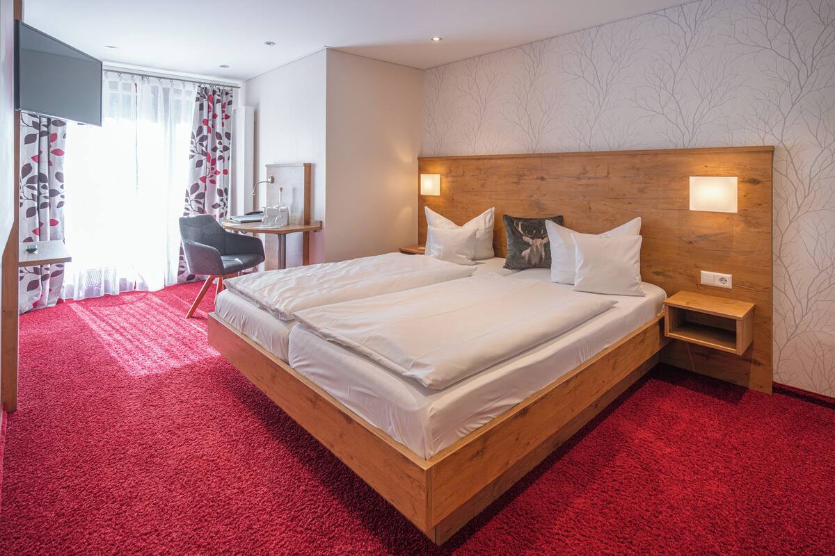 Ein Hotelzimmer mit einem hölzernen Bett, rotem Teppich, weißer Bettwäsche, einem Schreibtisch mit Stuhl und einem Fernseher.