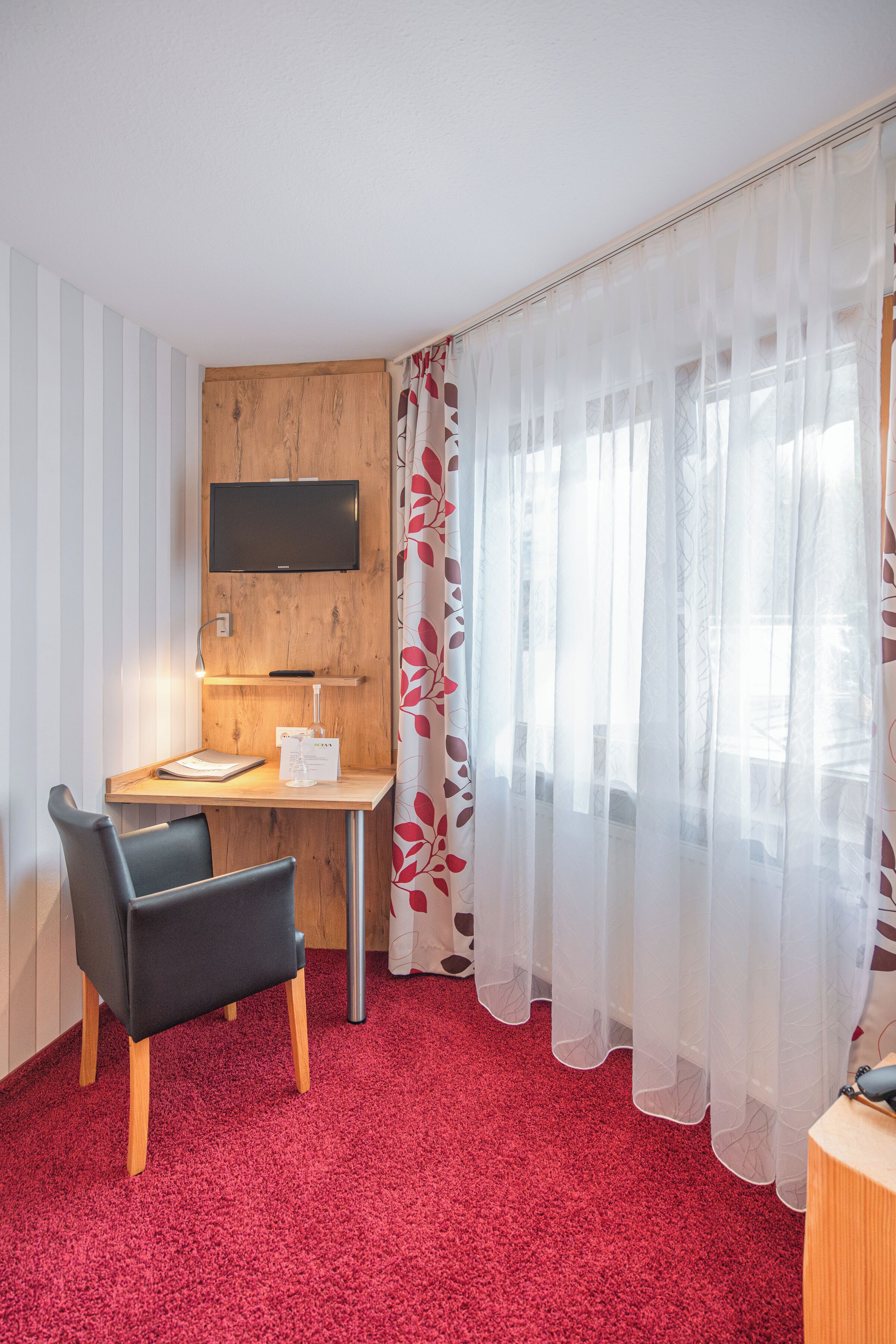 A hotel room with a wooden desk, a black chair, a TV mounted on the wall, and a red carpet.