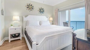 Une chambre à coucher avec un lit blanc, deux tables de chevet avec des lampes, un ventilateur de plafond et un balcon avec vue.