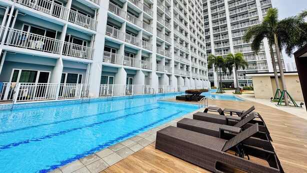 A large swimming pool with a wooden deck and lounge chairs, surrounded by a multi-story hotel building with balconies. - 180° Manila View near Airport, MOA, PICC and World Trade Center w/ Massage chair (Pasay City)