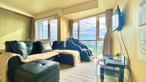 A modern living room with a sectional sofa, a massage chair, a glass coffee table, and a large window offering a view of the city. - 180° Manila View near Airport, MOA, PICC and World Trade Center w/ Massage chair (Pasay City)