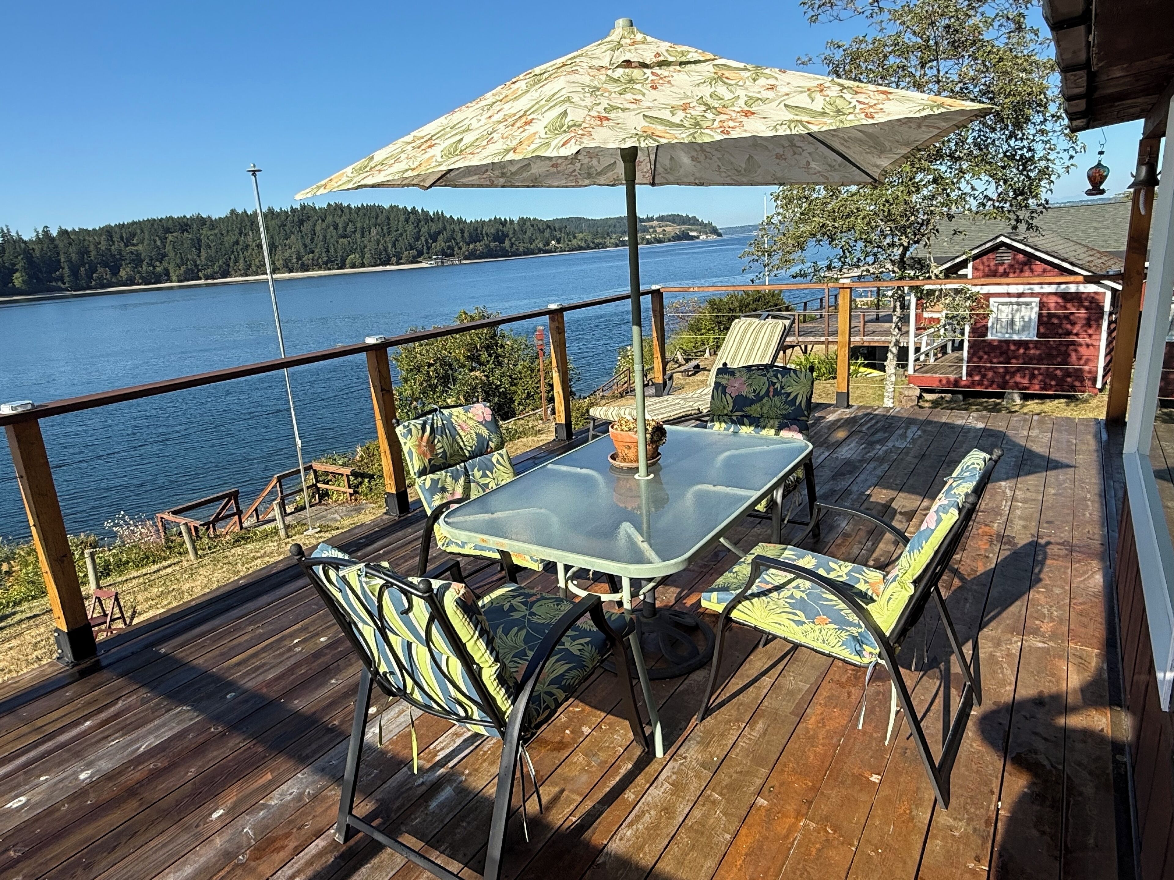 Une terrasse en bois avec une table et des chaises en verre, un parasol et une vue sur un lac.