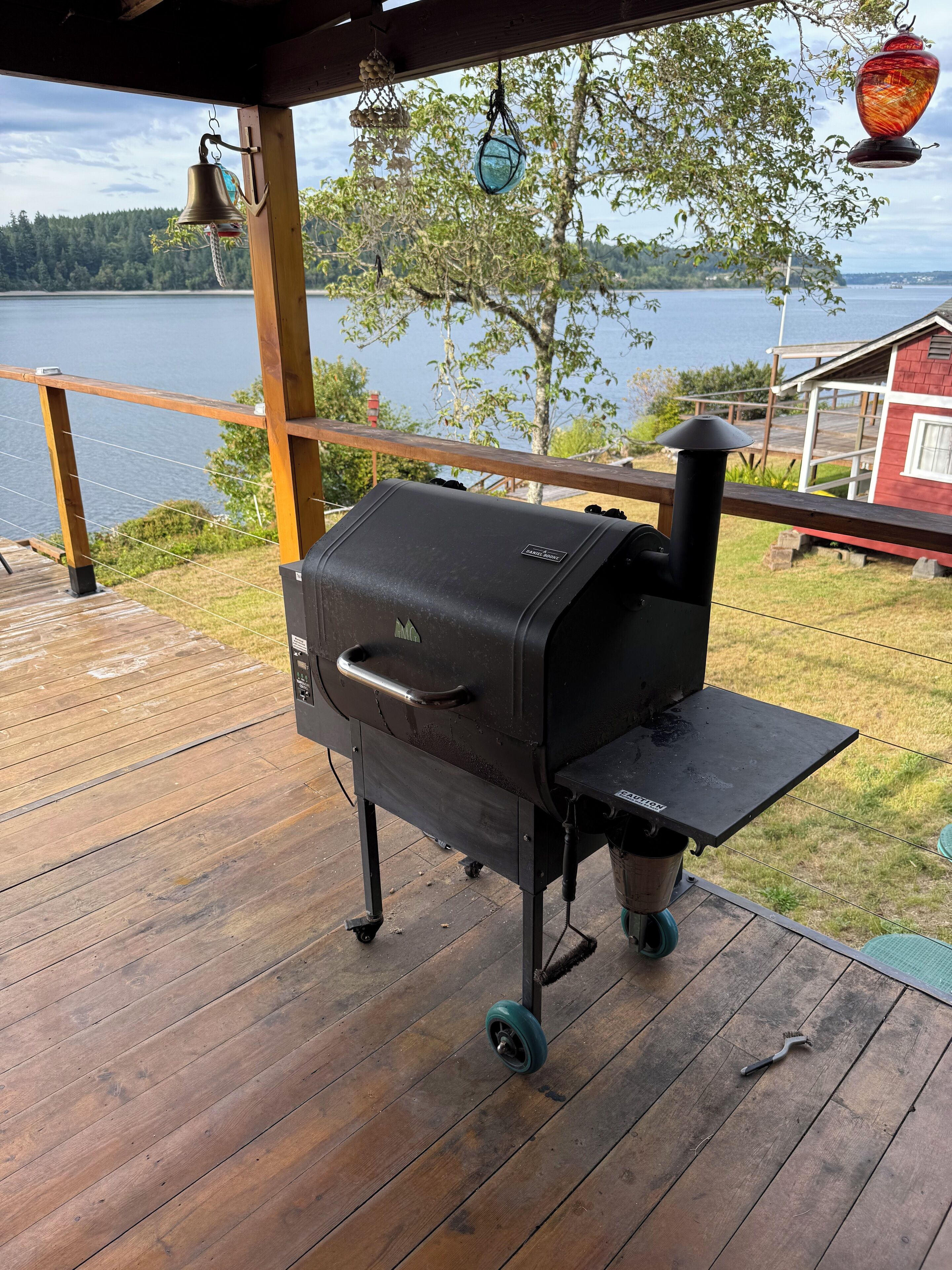 Ein Grill auf einer Holzterrasse mit Blick auf einen See und ein rotes Gebäude im Hintergrund.
