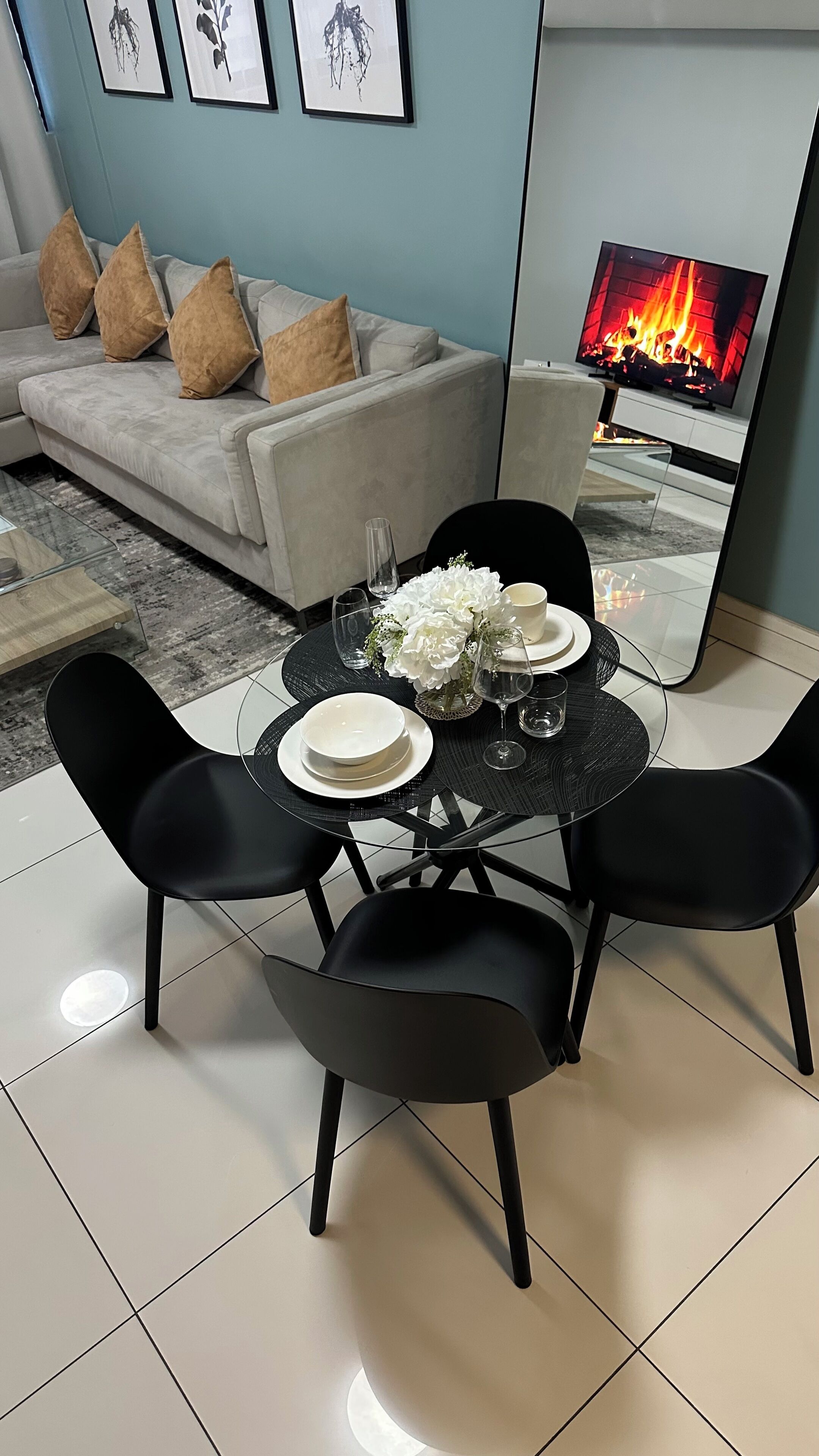 Un comedor con una mesa redonda dispuesta para dos personas, rodeada de sillas negras. En el fondo, un sofá con cojines color beige y un televisor de pantalla plana que muestra una chimenea.