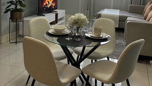 Una sala de estar moderna con una mesa redonda de vidrio, cuatro sillas, un televisor de pantalla plana mostrando una chimenea, un sofá y una ventana con cortinas.