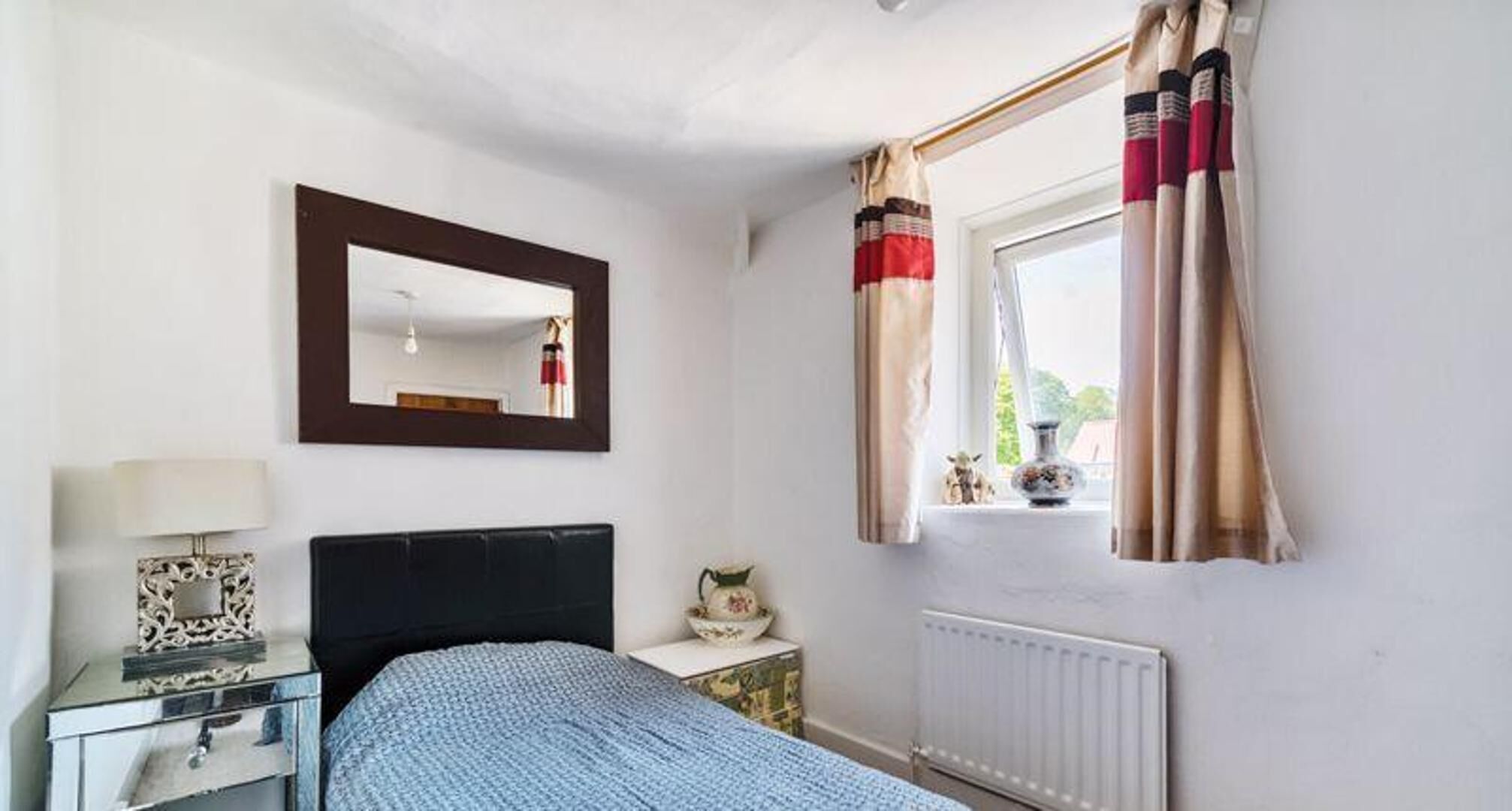 Une chambre à coucher comprenant un lit, une table de chevet, un miroir, une fenêtre avec des rideaux et un radiateur.