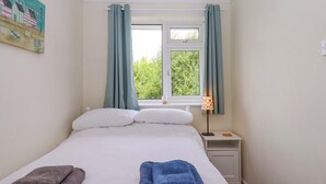 A neatly made bed with two pillows and two folded towels, a bedside lamp, and a window with curtains. - Hedgehogs (Barnstaple)