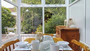 A conservatory with a table set for dining, surrounded by glass walls and a view of greenery. - Hedgehogs (Barnstaple)
