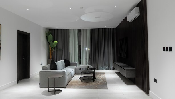 A modern living room with a grey sofa, a round coffee table, and a large window with curtains. - The VVS Apartments - Two-Bedroom Apartment with Pool View (Accra)