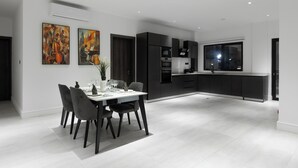 A modern kitchen with dark cabinetry, a dining area with a white table and chairs, and two abstract paintings on the wall. - The VVS Apartments - Two-Bedroom Apartment with Pool View (Accra)