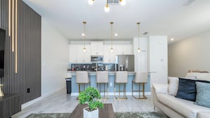 A modern kitchen with white cabinets, a central island, and bar stools. There is a dining area with a sofa and a coffee table. - Beautiful 4 BR Close to Disney (Kissimmee)