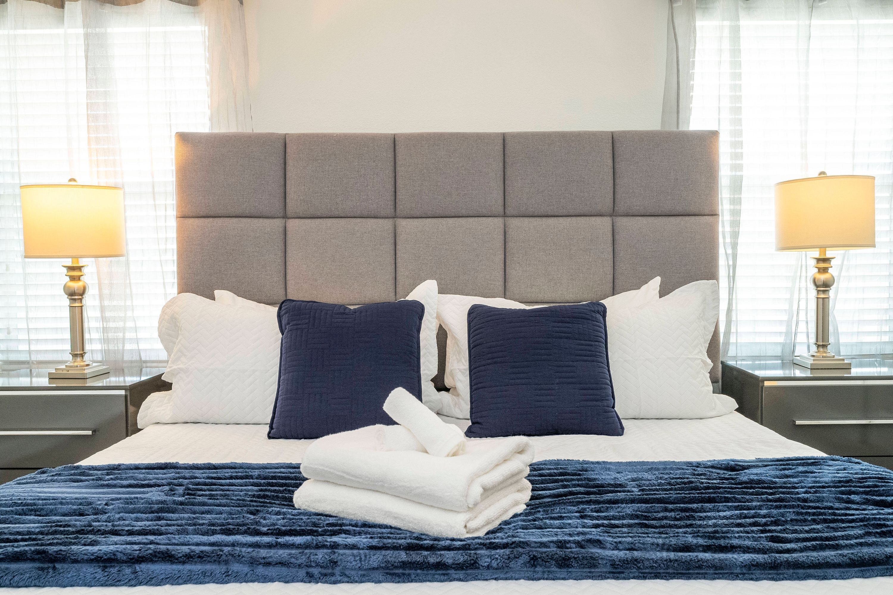 Een net opgemaakt bed met een grijs gestoffeerd hoofdbord, witte en marineblauwe kussens en een opgevouwen witte handdoek op een blauwe gebreide plaid.