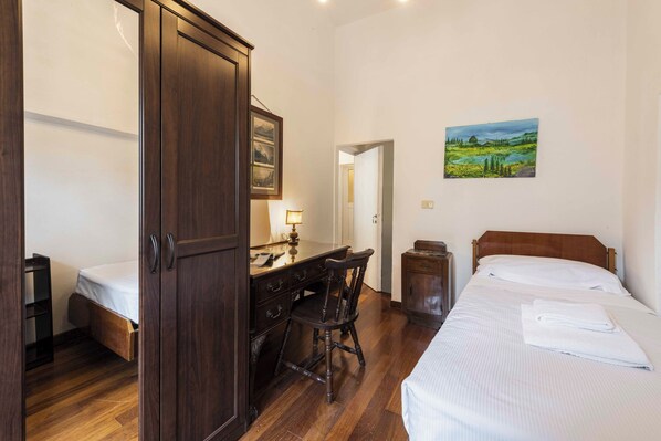 A bedroom with a wooden wardrobe, a desk with a chair, a bed with white linens, and a landscape painting on the wall.