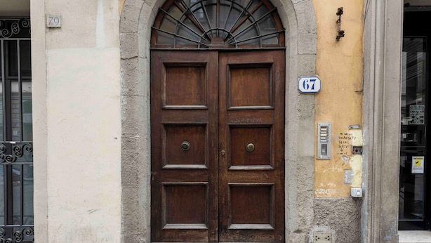 A double wooden door with a small window above it, a number '67' sign, and a wall with a rough texture and a utility box. - Ghibellina Retreat by MMega (Florence)