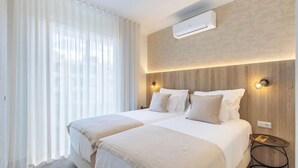 A hotel room with two beds, a large window with sheer curtains, an air conditioning unit, and a small round table with a lamp. - Vila Baleira Residence (Funchal)