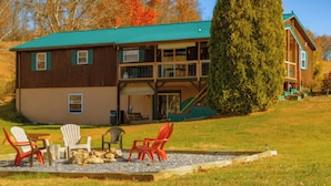 A two-story lodge with a green roof, outdoor seating area with Adirondack chairs, and a large tree in the foreground.
