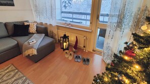 A cozy living room with a grey sofa, a small table with decorative items, a radiator, and a window with a view of snow-covered trees. - Spacious apartment with balcony and free WiFi (Rovaniemi)