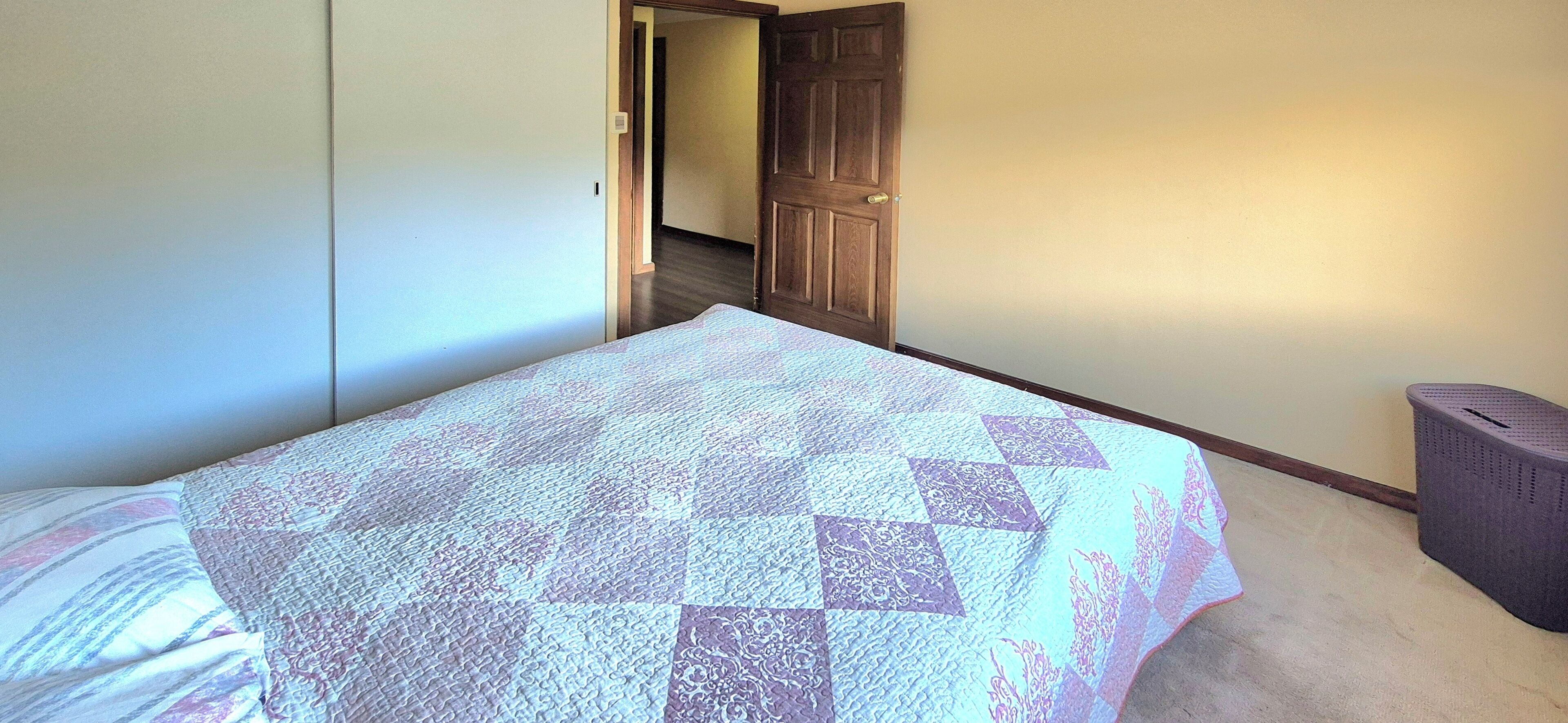 A bedroom with a bed covered in a patterned quilt, a wardrobe, a door leading to another room, and a laundry basket.