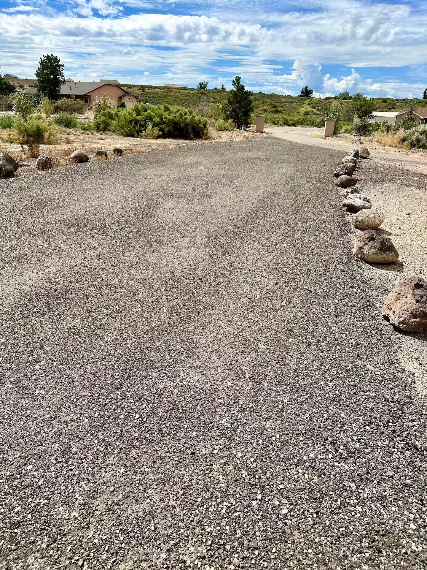 A gravel road with rocks on the side, surrounded by sparse vegetation and a few buildings in the background.