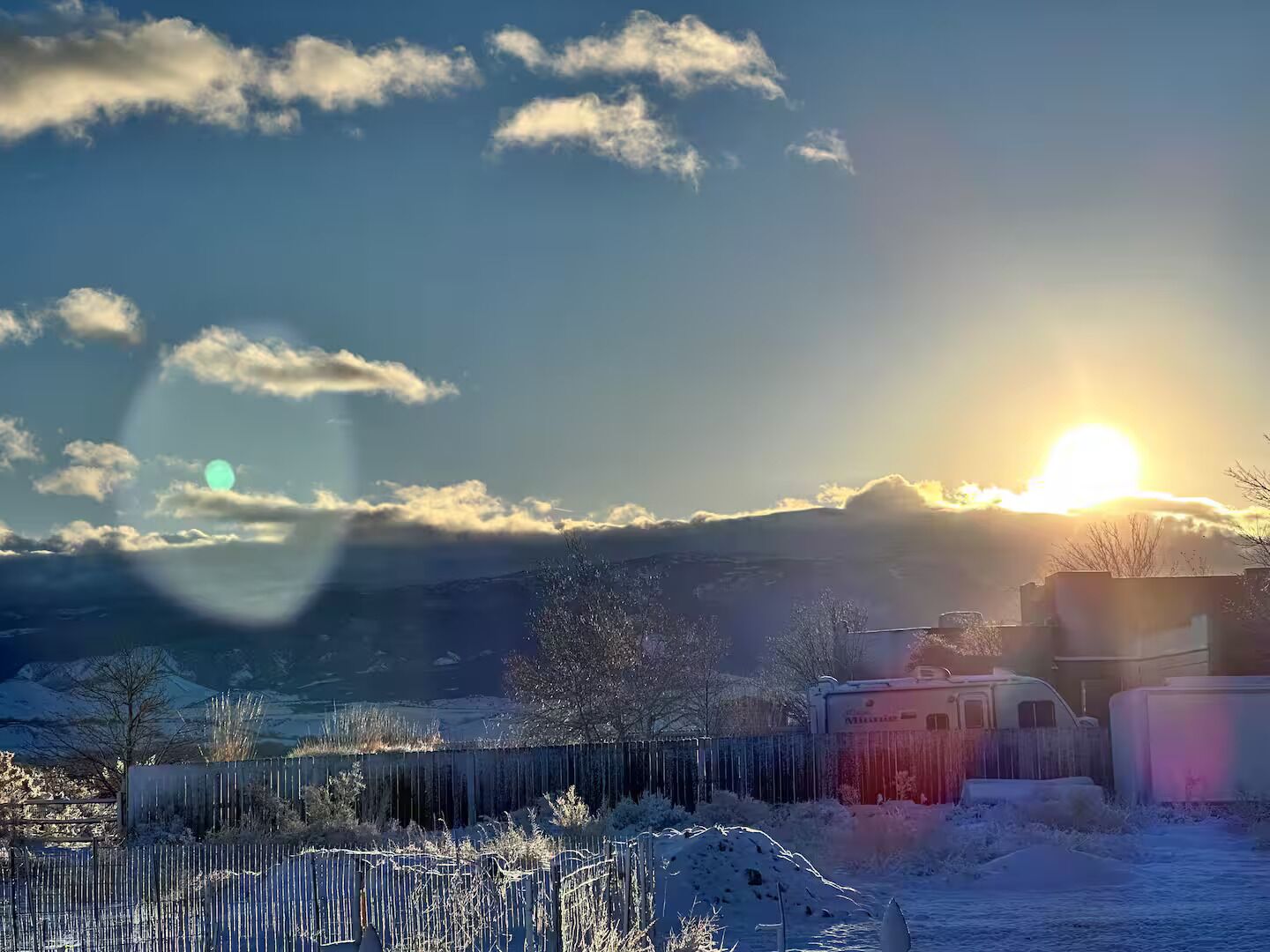 A snowy landscape with a sun setting behind a mountain range, a fence, and a trailer.