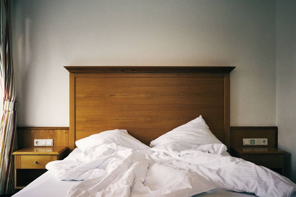A hotel room with a wooden headboard, a single bed with white linens, two bedside tables with clocks, and a power outlet on the left table.