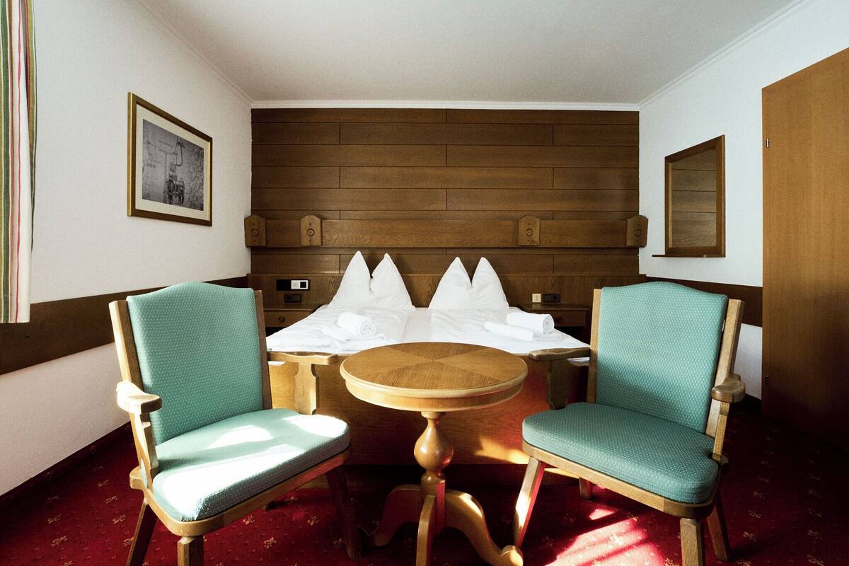 A hotel room with a wooden headboard, two green upholstered chairs, a round wooden table, and a bed with white linens.