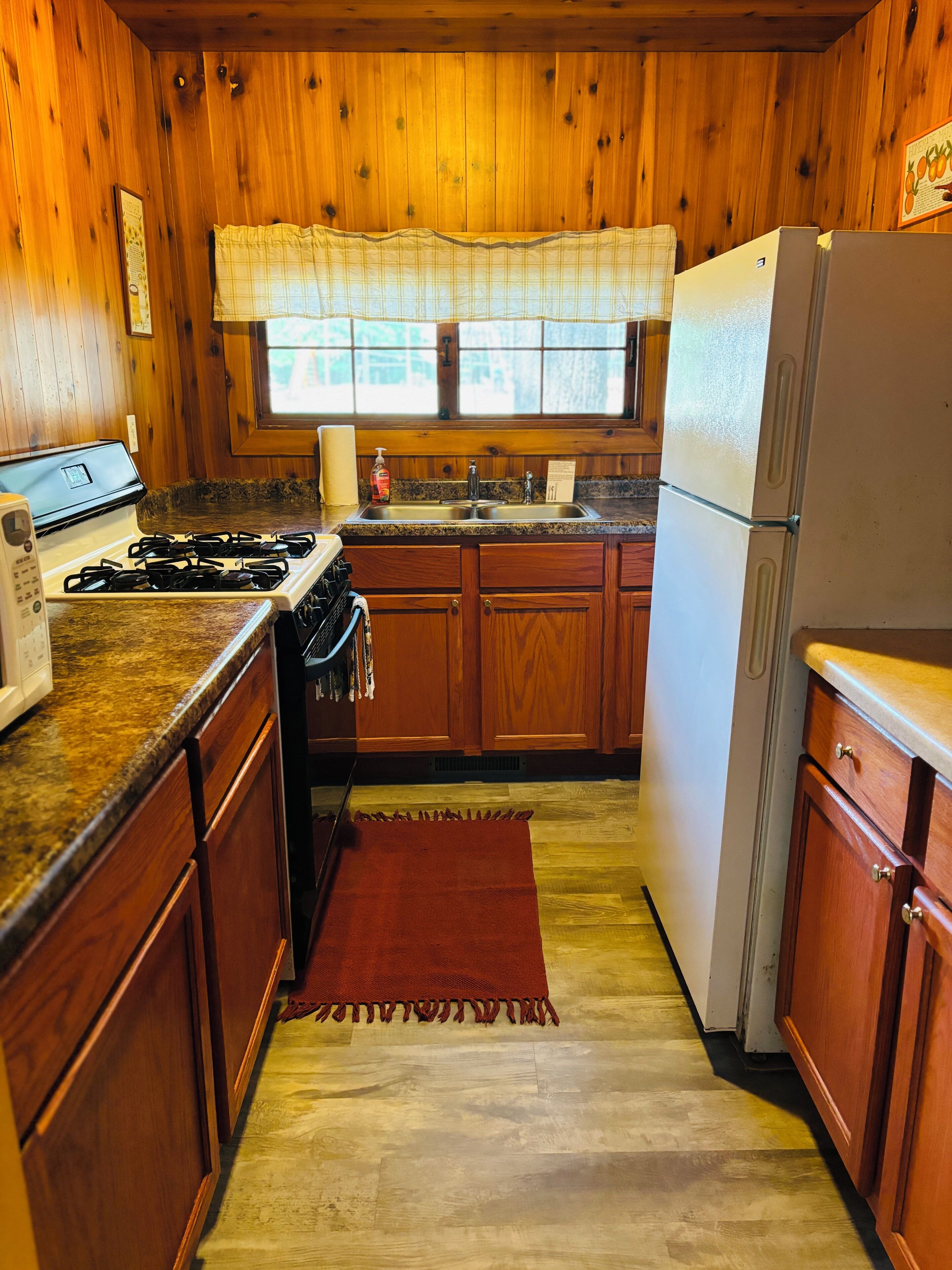 A compact kitchen with wooden cabinets, a gas stove, a refrigerator, and a window with a plaid valance.