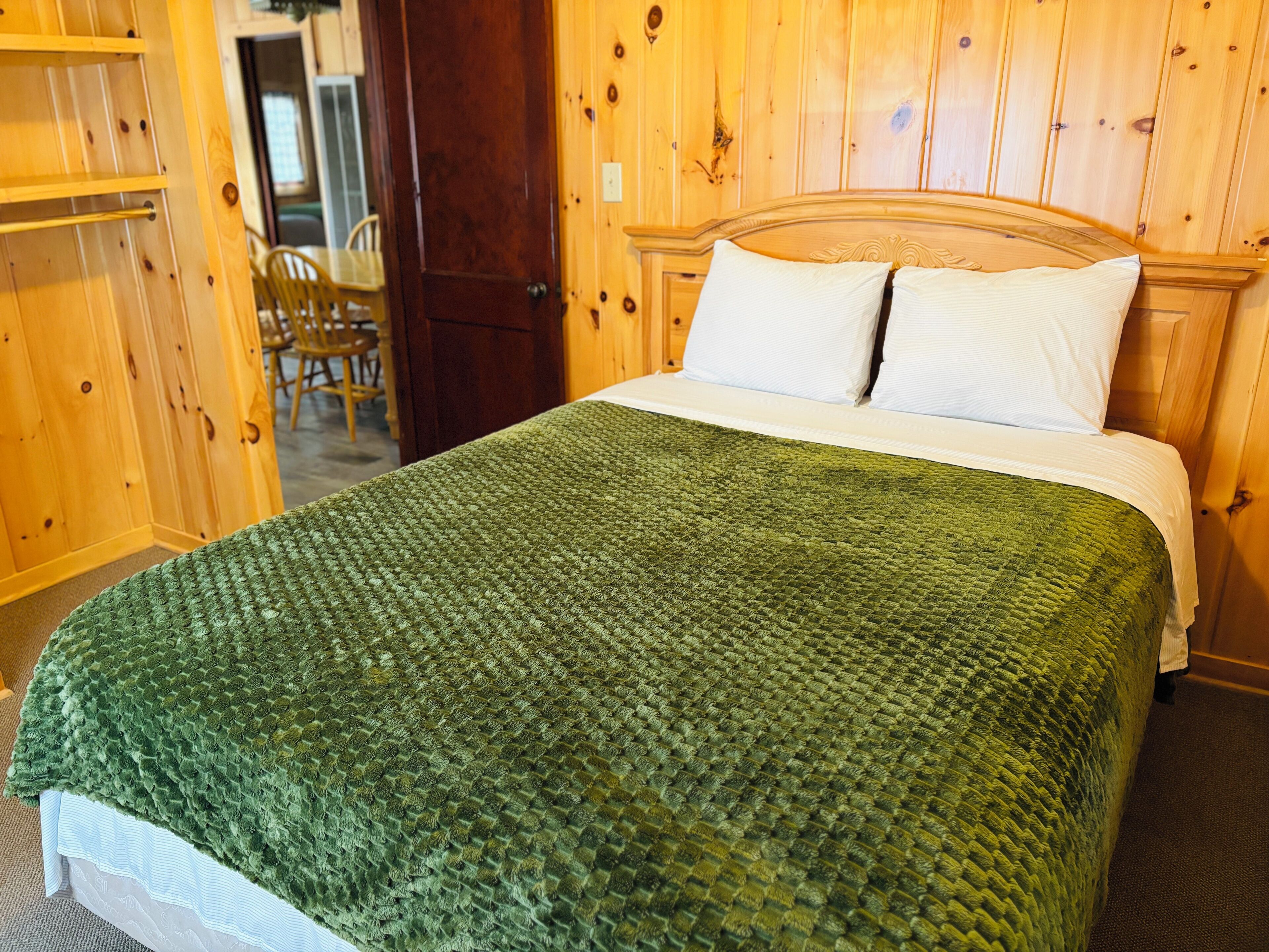 A bed with a green quilt, white pillows, and a wooden headboard in a room with wooden walls and a door.