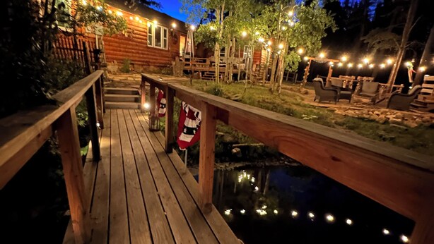 A wooden deck leading to a cabin with string lights, a small pond, and outdoor furniture.