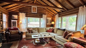 A cozy log cabin interior with a wooden ceiling, a large sofa, a coffee table, and a patterned rug.