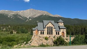 A stone church with a bell tower, set against a backdrop of mountains and clear blue sky.