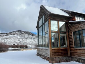 Una cabaña de madera con amplios ventanales, ubicada frente a un paisaje nevado con una montaña al fondo.