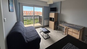 Un espacio habitable compacto con sofá, una mesita, kitchenette y un balcón con vistas a edificios.