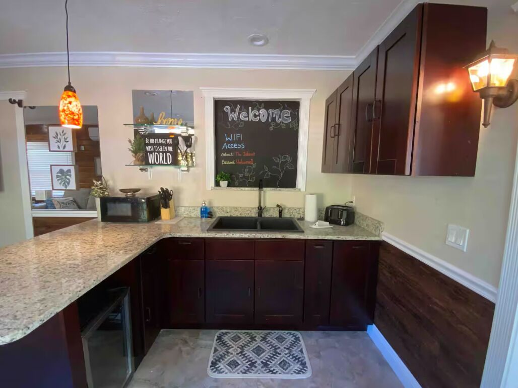 Una cocina con armarios de madera oscura, encimera de granito, una pizarra con 'Welcome' y 'WiFi Access' escrito y una lámpara colgante.