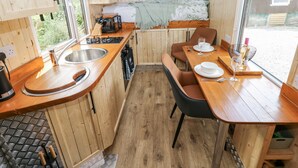 A compact kitchen with wooden cabinets, a sink, a stove, and a dining area with a table and chairs. - The Hamper (Carmarthen)