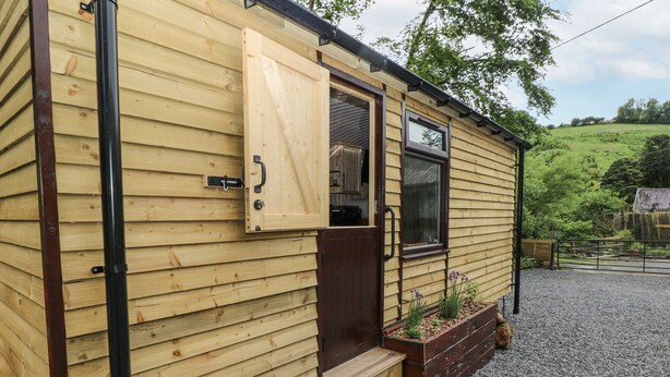 A wooden cabin with a sliding door, a window, and a small outdoor seating area with steps leading up to it. - The Hamper (Carmarthen)